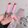 Two pink lipsticks with heart-shaped caps held by a hand, with a clear box labeled 'Gee Be' in the background.