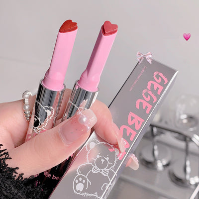 Two pink lipsticks with heart-shaped caps held by a hand, with a clear box labeled 'Gee Be' in the background.