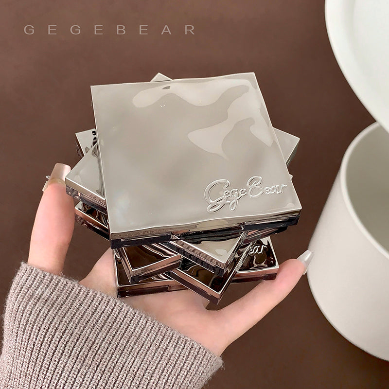 Stack of metallic square items with 'Gegebear' branding held in a hand against a brown background.