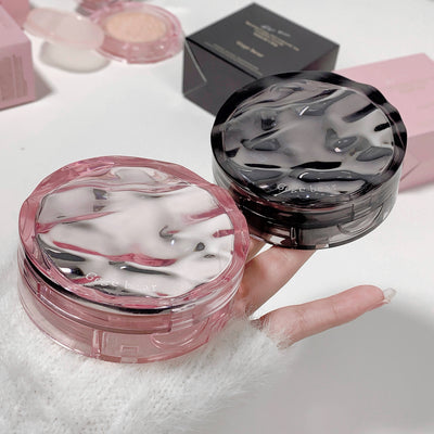 Two compact powder cases, one pink and one black, held by a hand on a white surface.