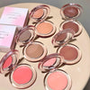 Set of makeup blushes in various colors on a round surface with a pink box in the background.