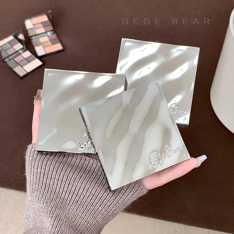 Two silver compact mirrors with a brand logo held by a hand on a brown surface.