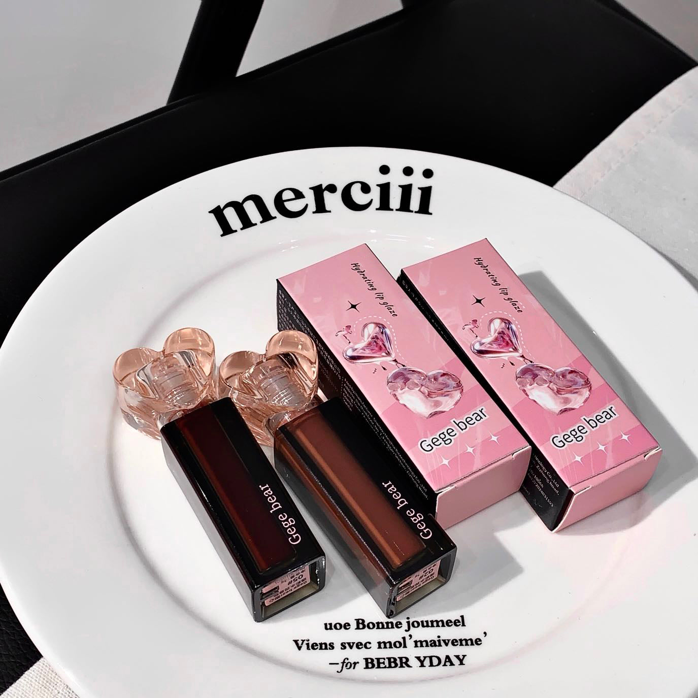 Two lip glosses and their packaging on a white plate with 'merciii' branding.