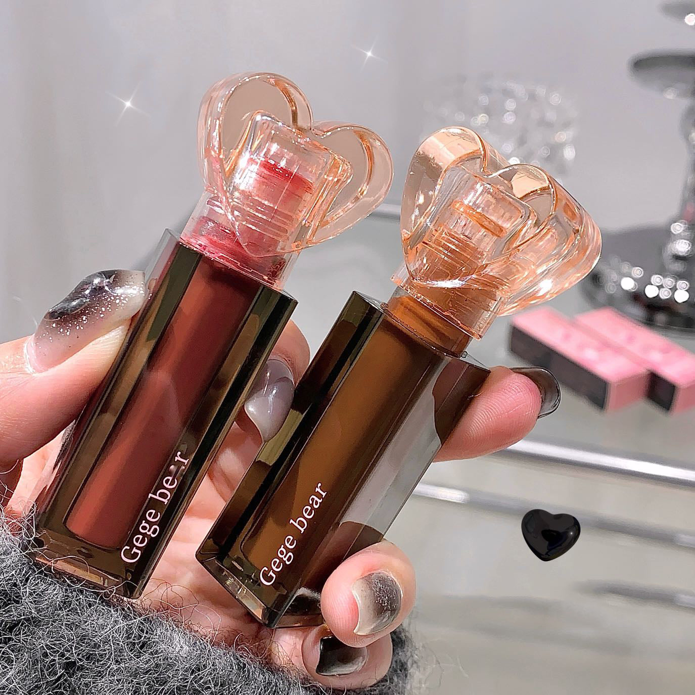 Two lip glosses with heart-shaped caps held by a hand against a blurred background.