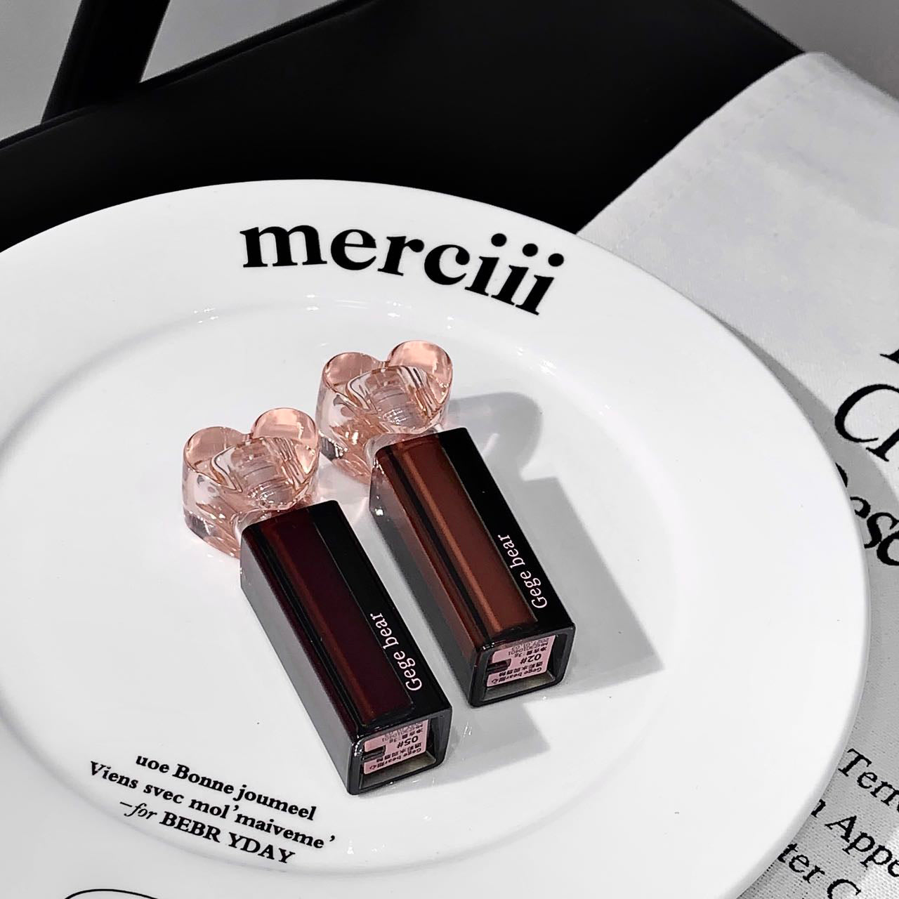 Two lipsticks with heart-shaped caps on a white plate with 'merciii' text.
