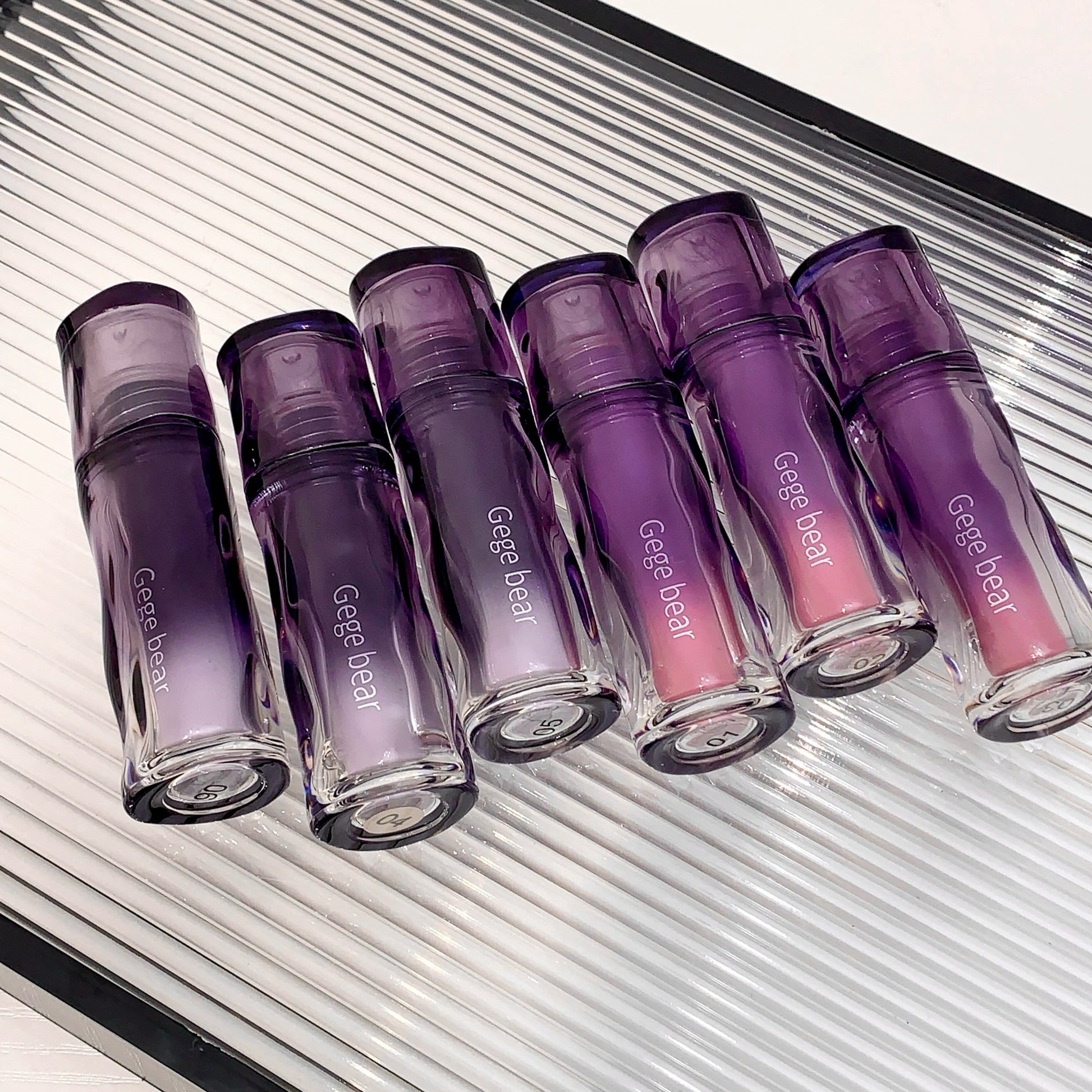 Set of purple and pink lip glosses with 'Geege bear' branding on a metallic surface.