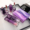 Purple cosmetic products including lipsticks and a book titled 'The She Book' on a reflective surface.