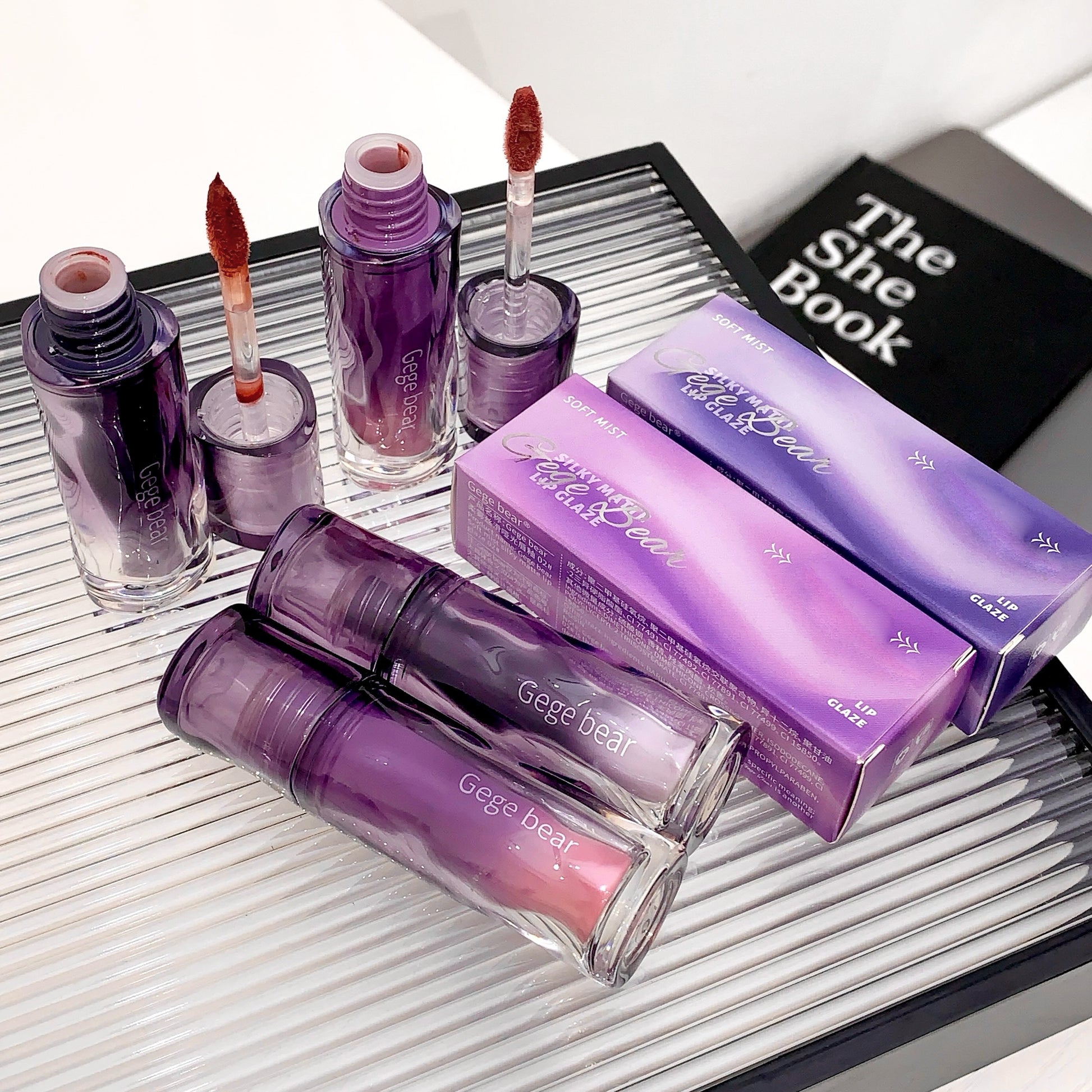 Purple cosmetic products including lipsticks and a book titled 'The She Book' on a reflective surface.