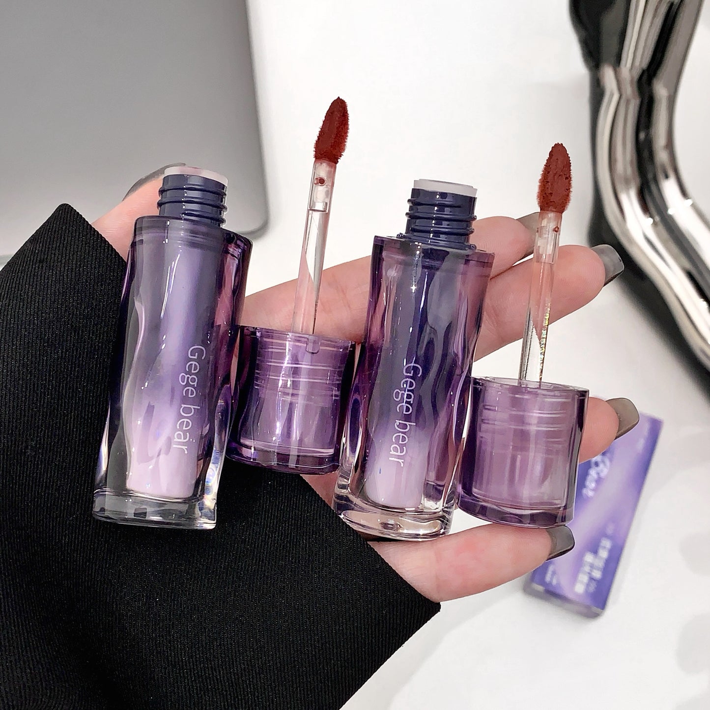 Hand holding small purple cosmetic bottles with brushes, branded 'Gege bear', against a neutral background.