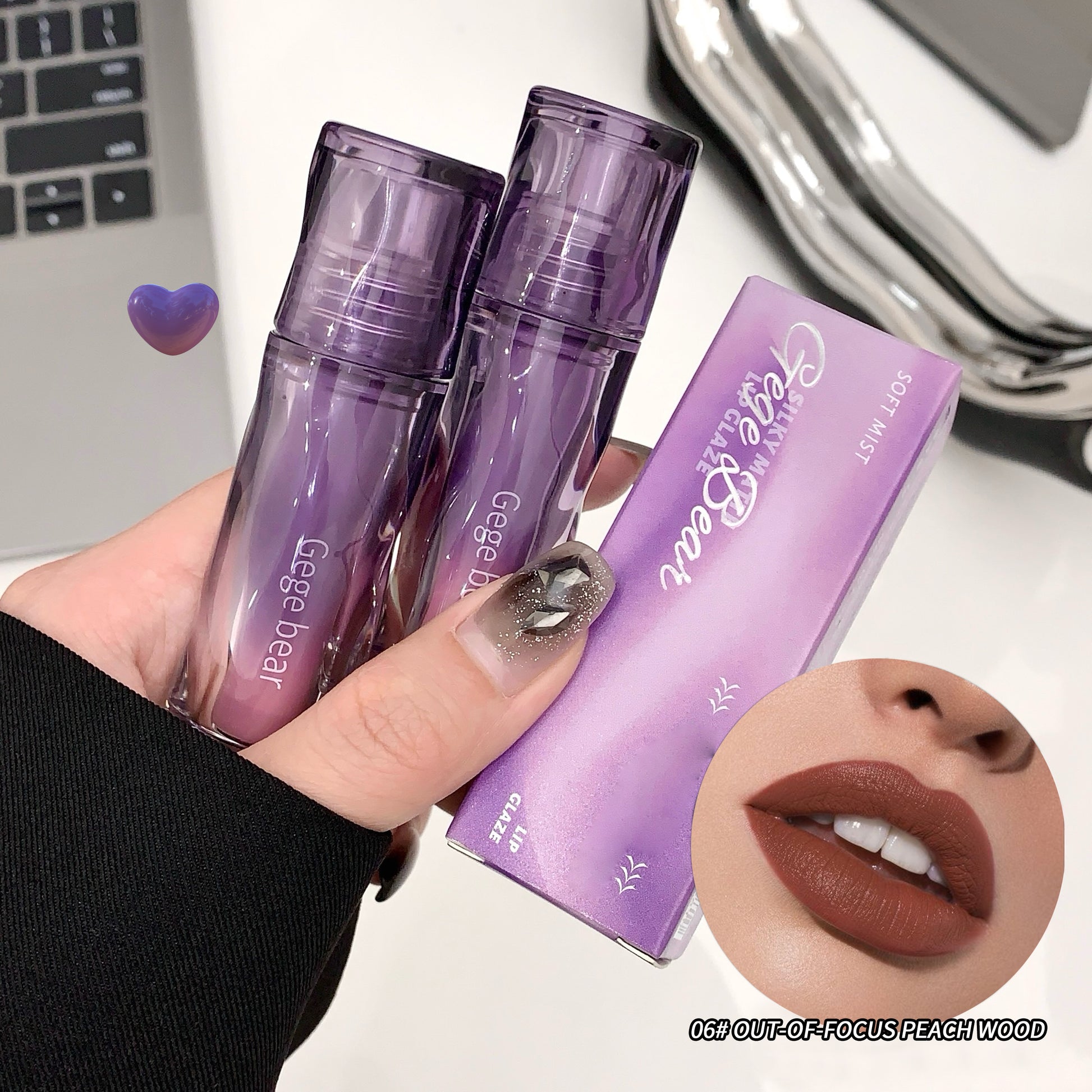 Purple lip gloss with packaging held by a hand, laptop, and mirror in the background.