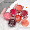 Hand holding small containers of lip gloss with a close-up of a lip gloss sample on a white surface.