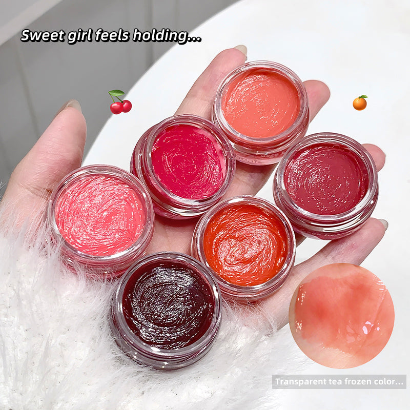 Hand holding small containers of lip gloss with a close-up of a lip gloss sample on a white surface.