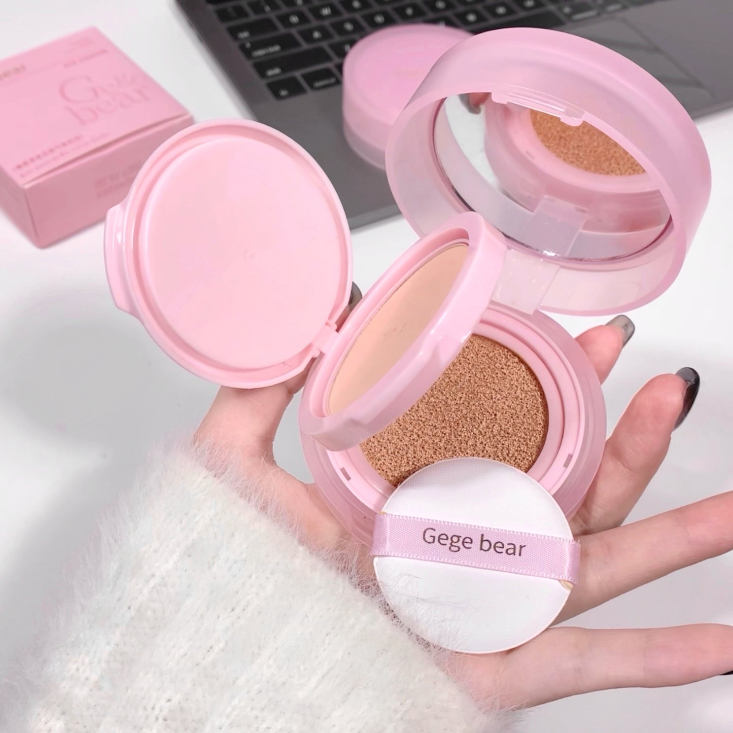 Pink compact powder with a sponge puff held by a hand, with a laptop in the background.
