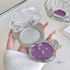 Compact powder case with silver and purple shades held by a hand on a light background