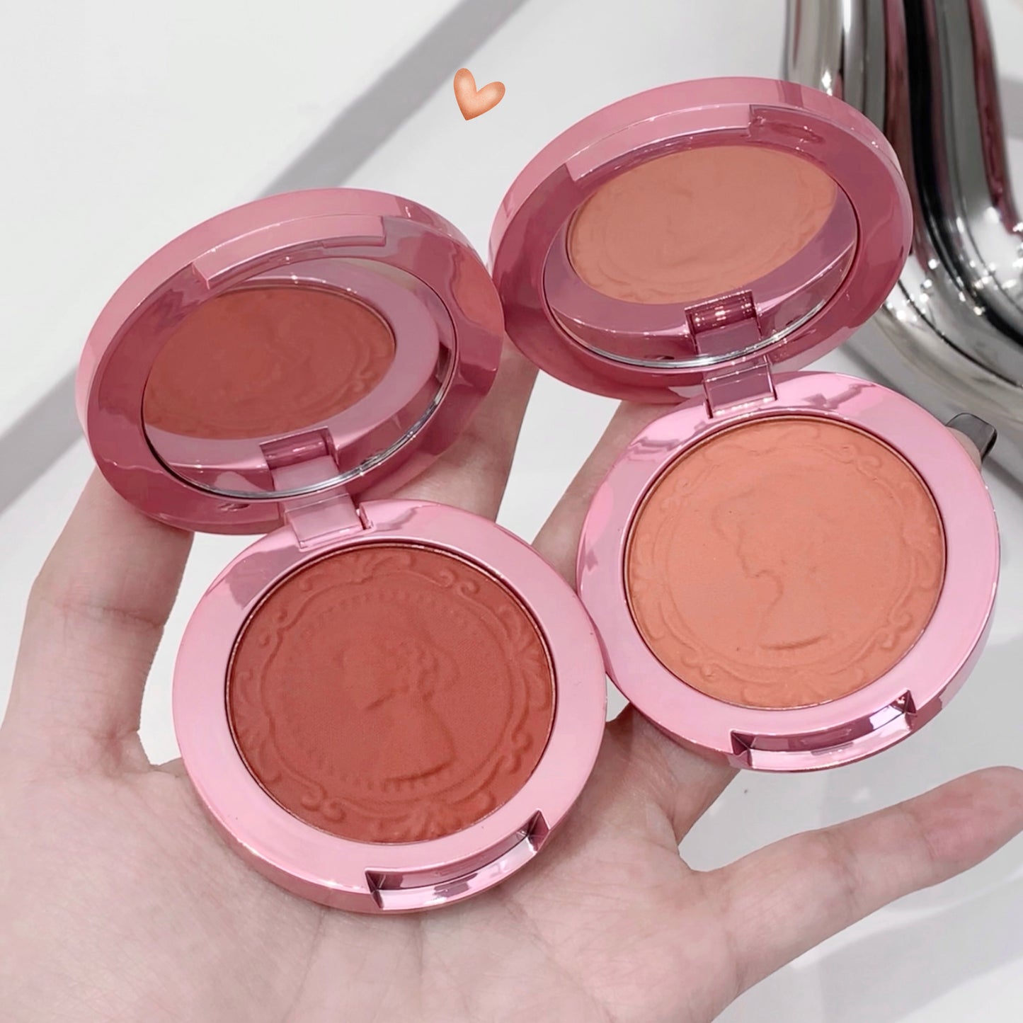 Four pink makeup blushes in open cases held in a hand with a white background.