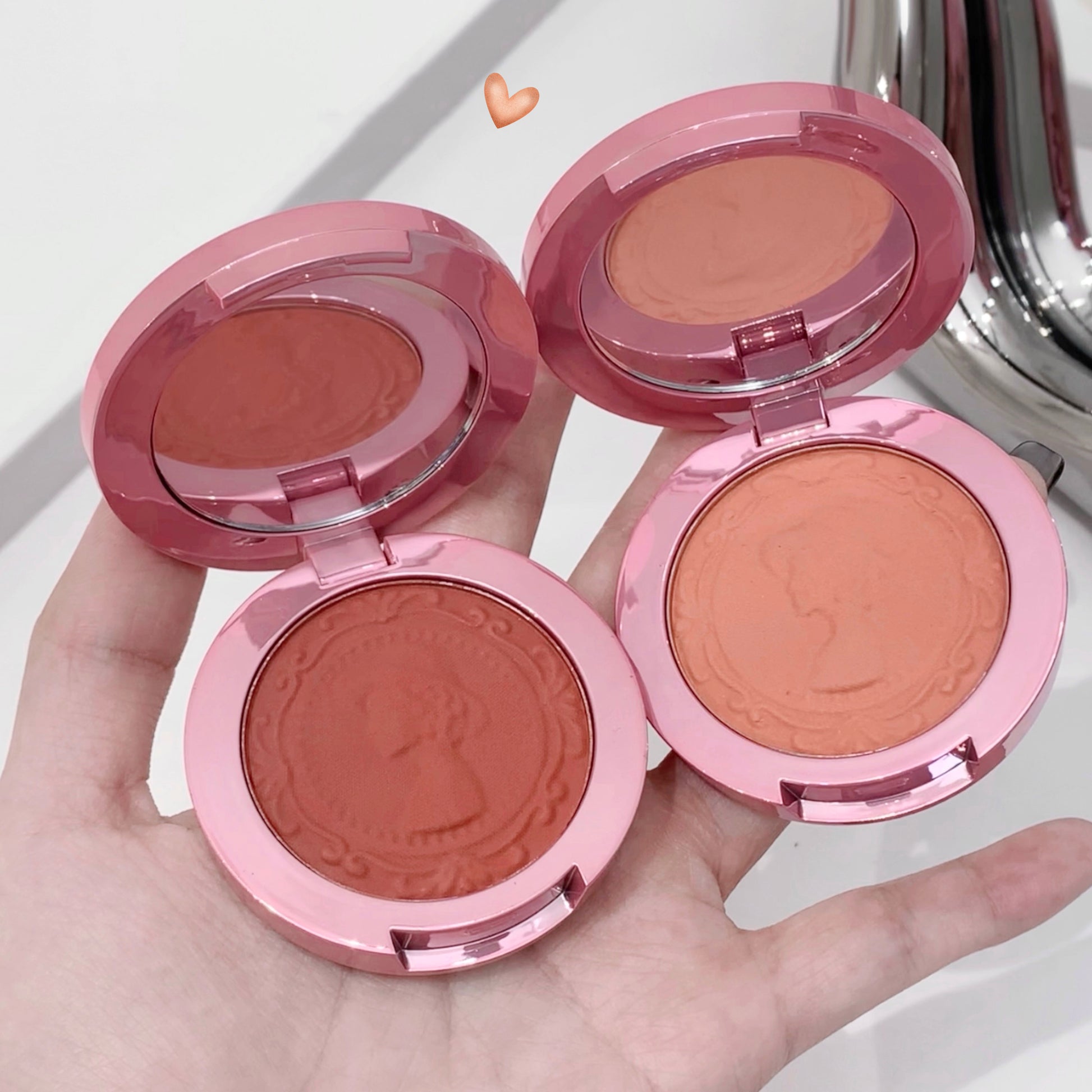 Four pink makeup blushes in open cases held in a hand with a white background.