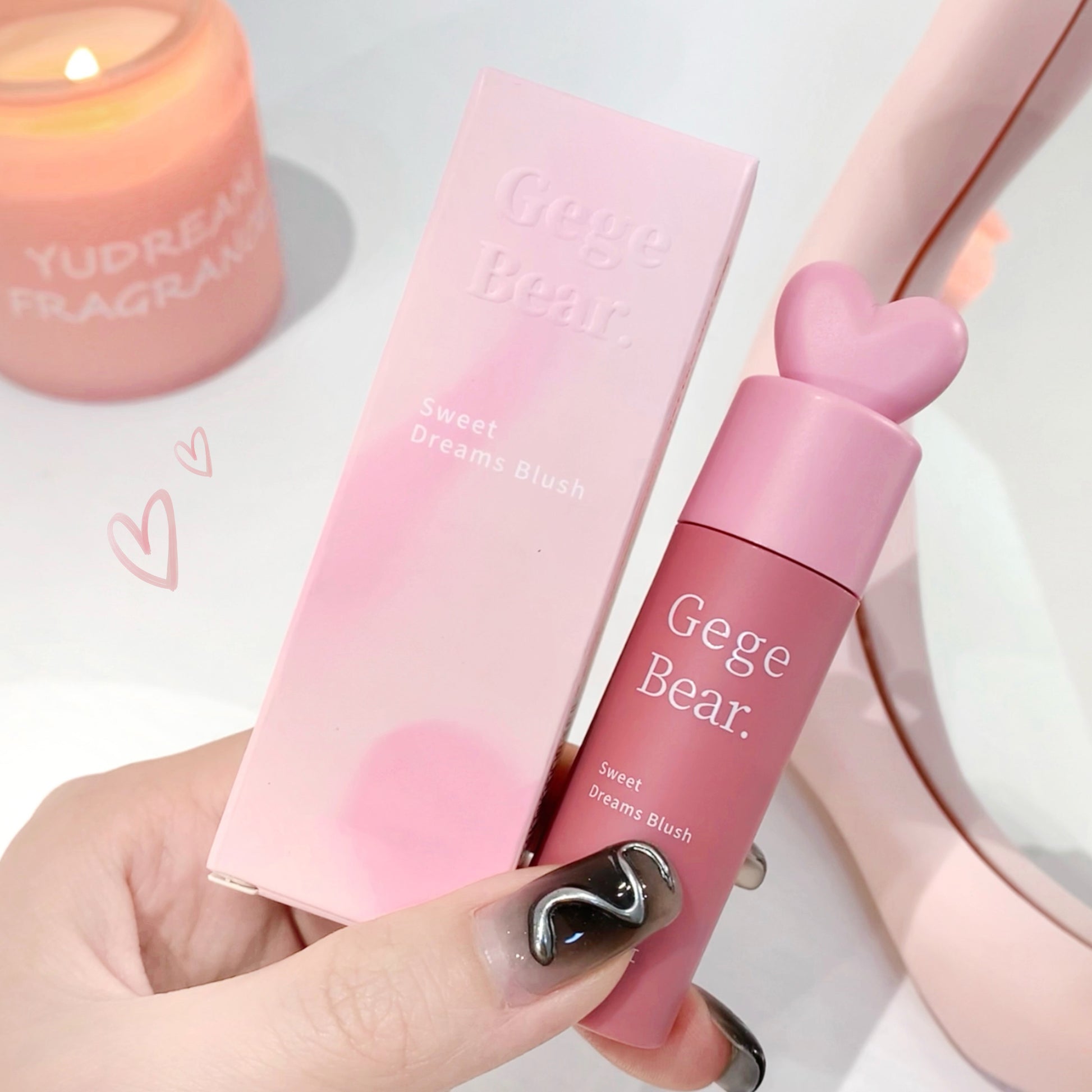 Pink cosmetic product with 'Gege Bear' branding held by a hand, next to a pink box and candle.