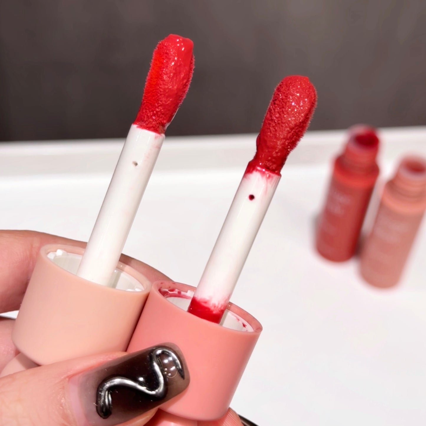 Two red lip glosses with white applicators held by a hand with black nail art on a white surface.