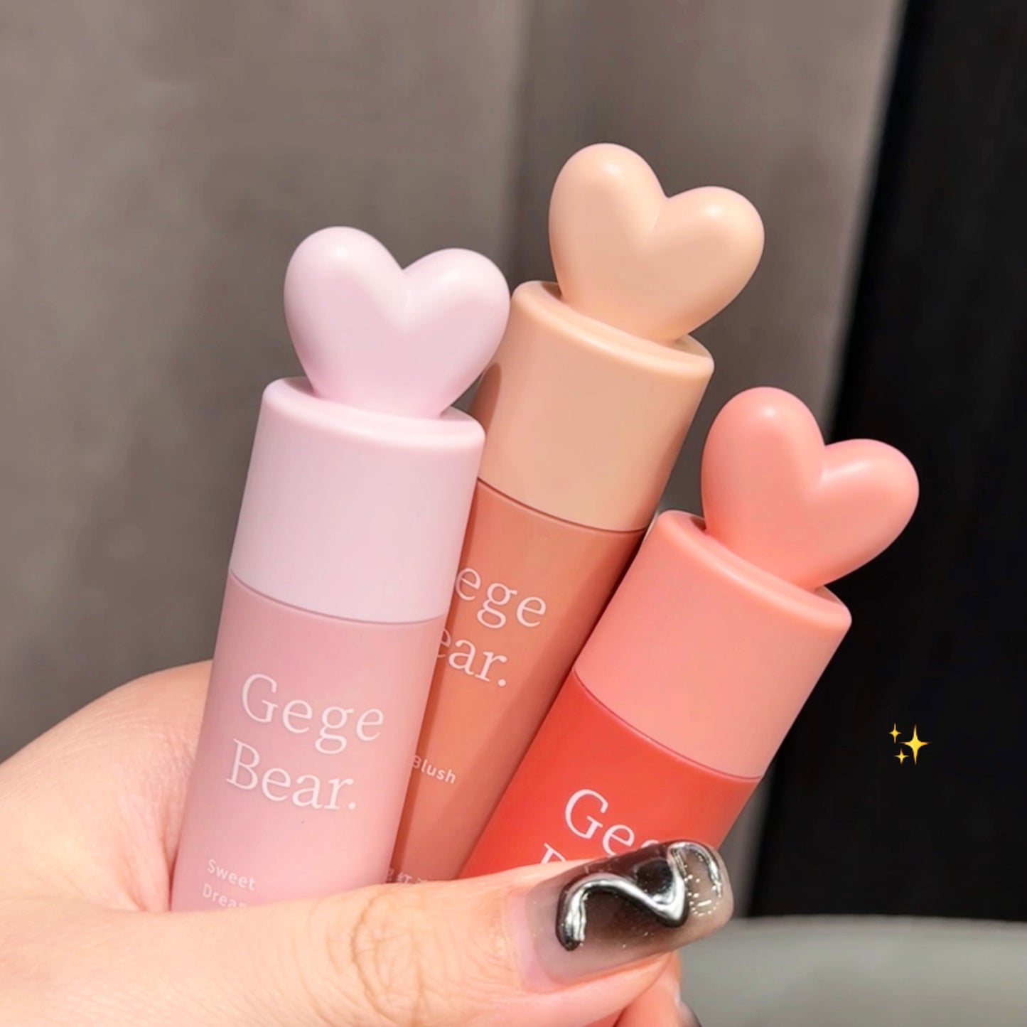 Three lip glosses with heart-shaped caps held by a hand, branded 'Gege Bear'.