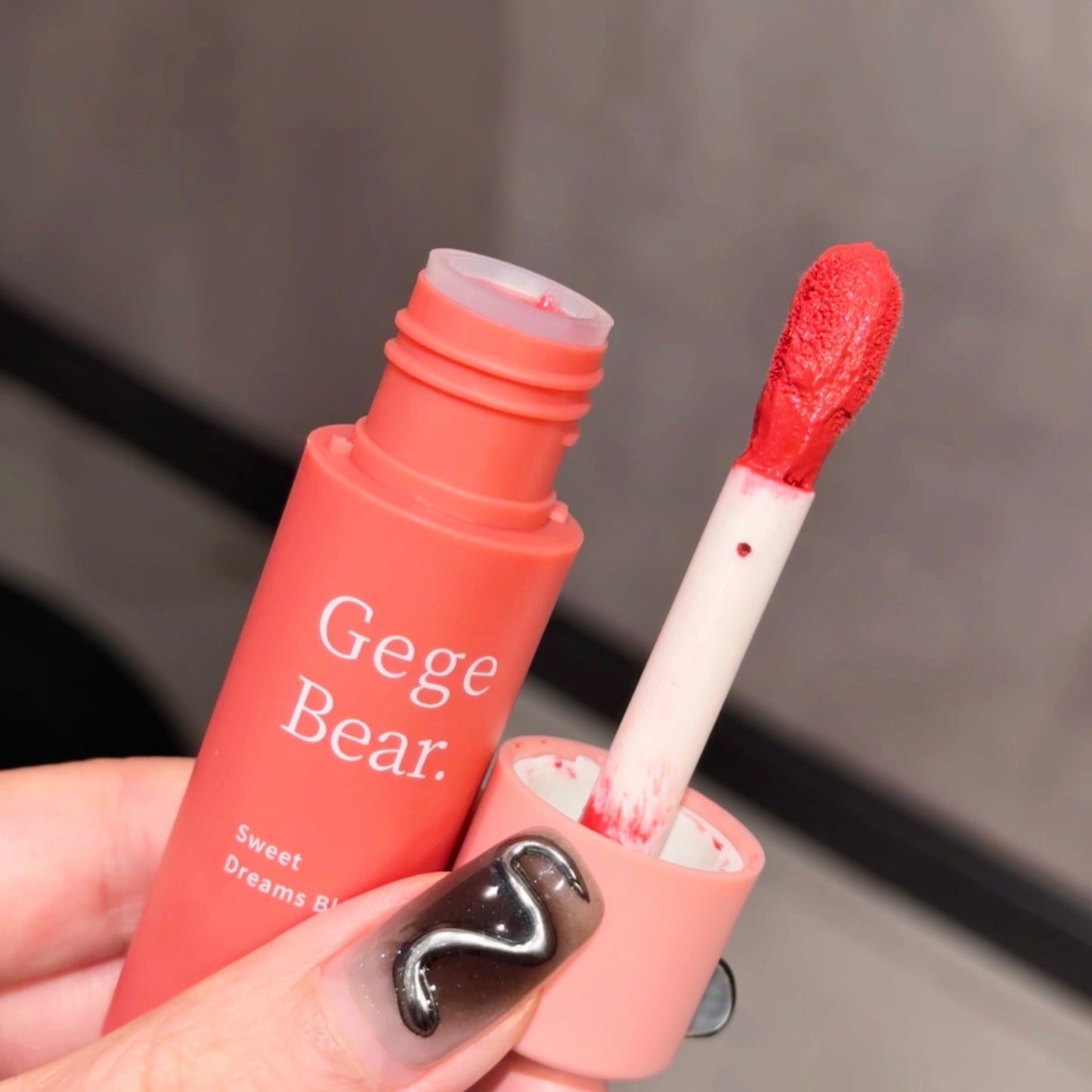 Red lip gloss with applicator held by a hand, brand 'Gege Bear' visible