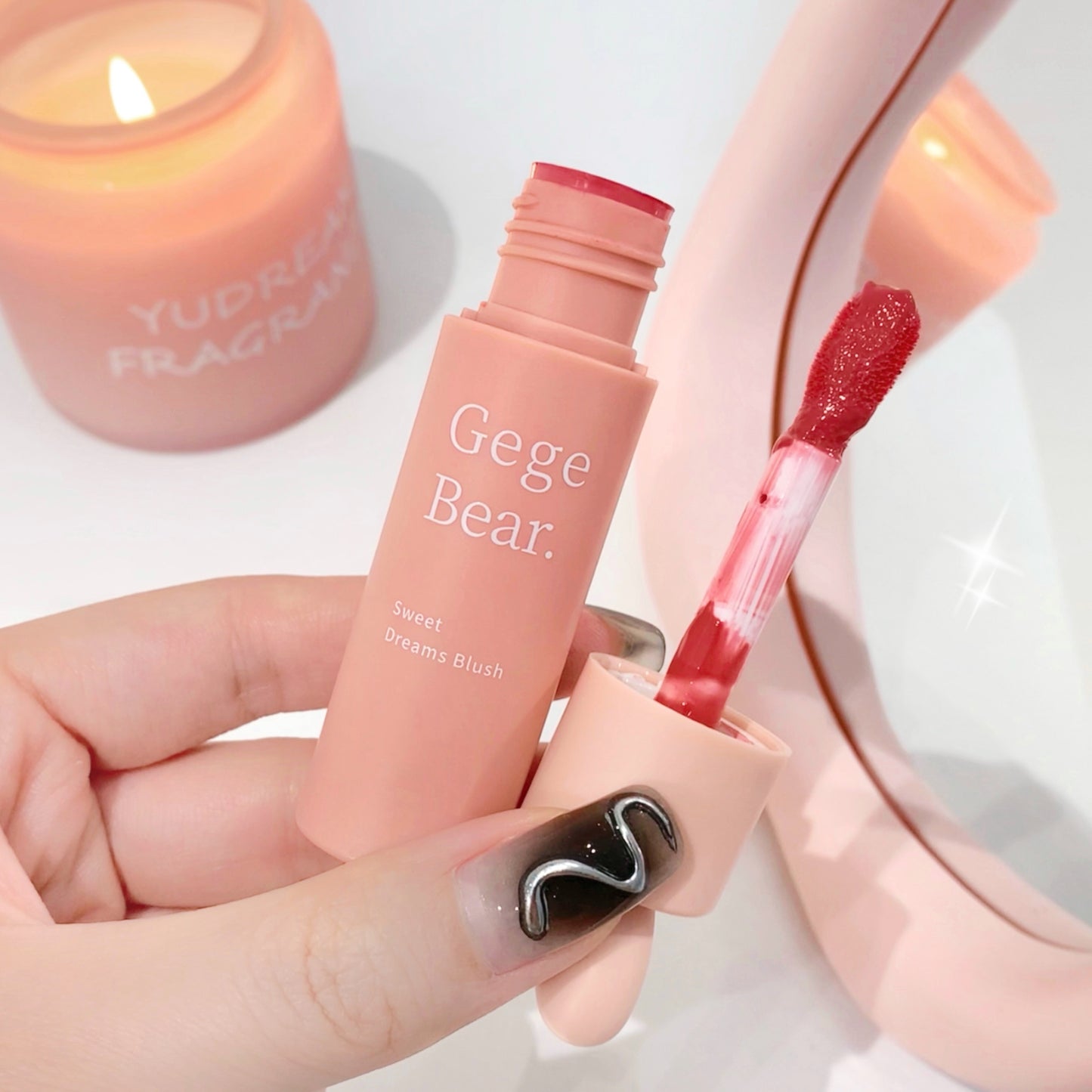 Hand holding a pink makeup product with 'Gege Bear' branding, blurred candle in the background