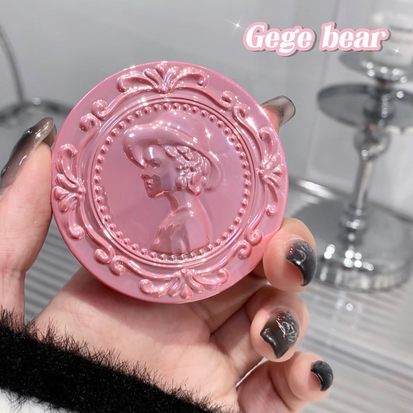 Pink decorative compact mirror held by a hand with black nail polish on a neutral background.