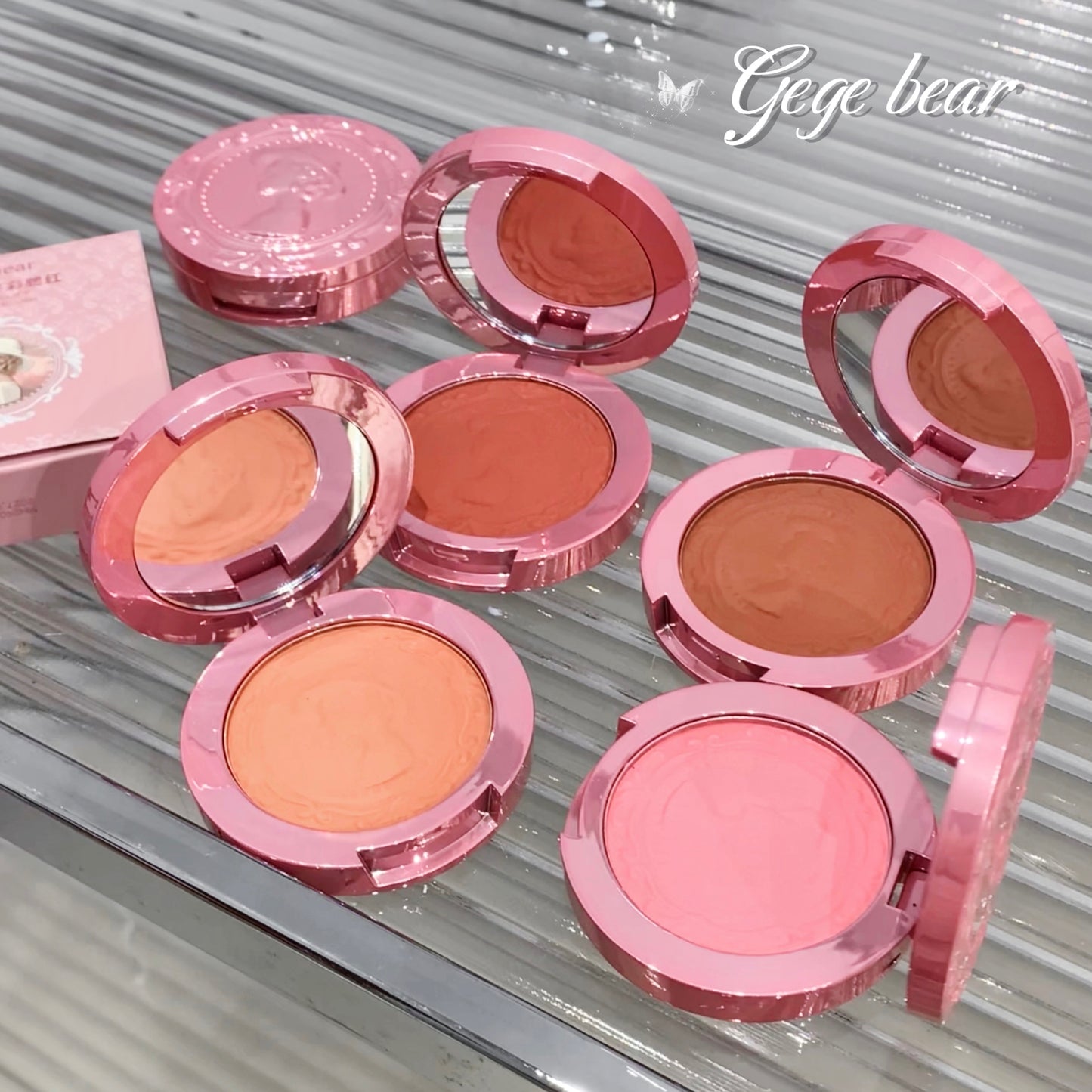 Pink makeup compacts with different shades of blush on a metallic surface, branded 'Geebear'.