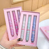 Set of four lip balms in pink packaging held by a hand on a light wooden surface.