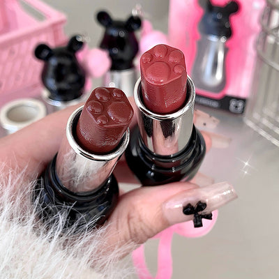Two lipsticks with paw-shaped caps held by a hand against a blurred background.