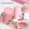 Pink lip balm with cherry design held by a hand, close-up of lips with product applied.
