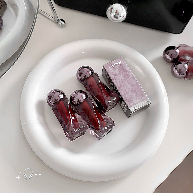 Three purple lip glosses and their packaging on a white plate with a blurred background.
