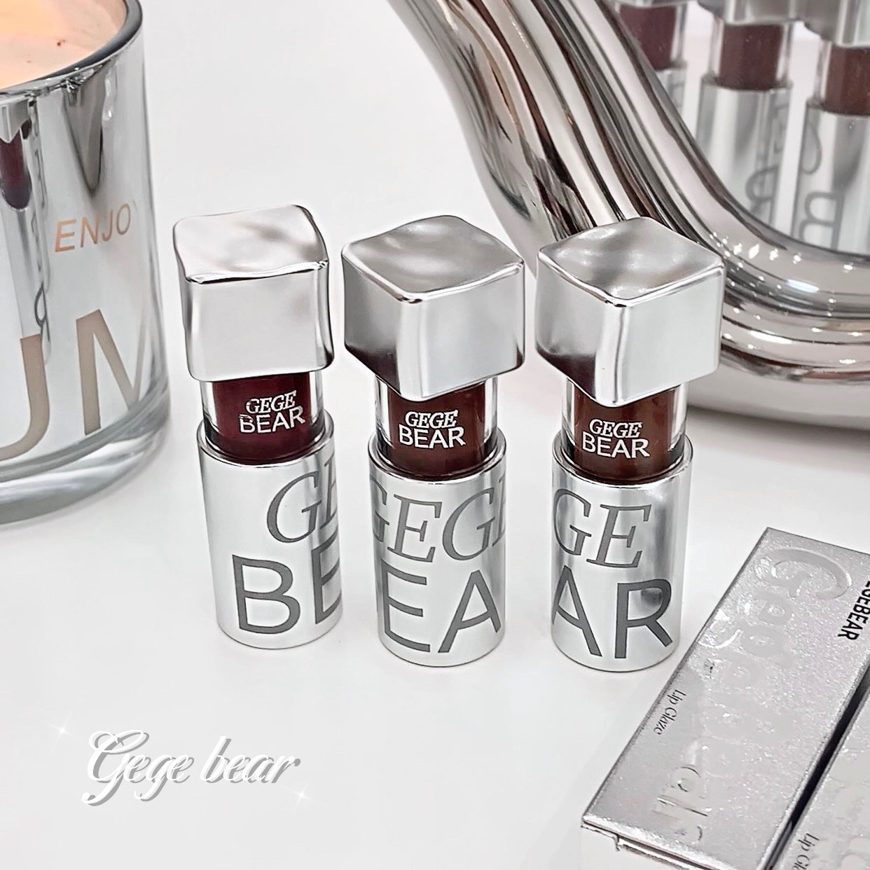 Three Gege Bear lip glosses with silver caps on a white surface.