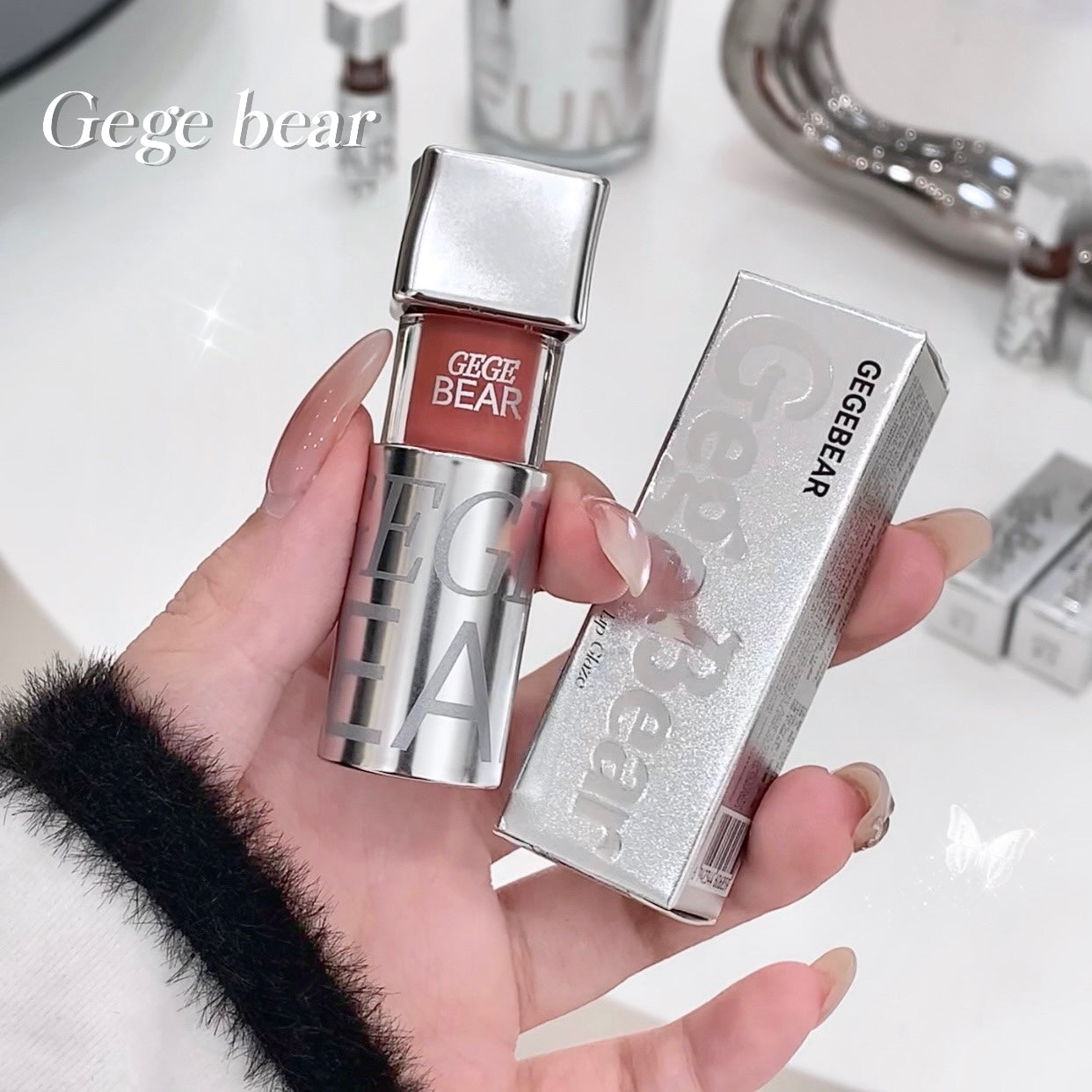 Hand holding a silver and red lip gloss with 'Gege Bear' branding against a blurred bathroom background.
