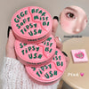 Pink compact mirror with green text held by a hand, close-up of a person's eye, and pink product on a beige surface.