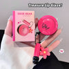 Pink lip gloss with floral design held by a hand, next to its packaging, on a neutral background.