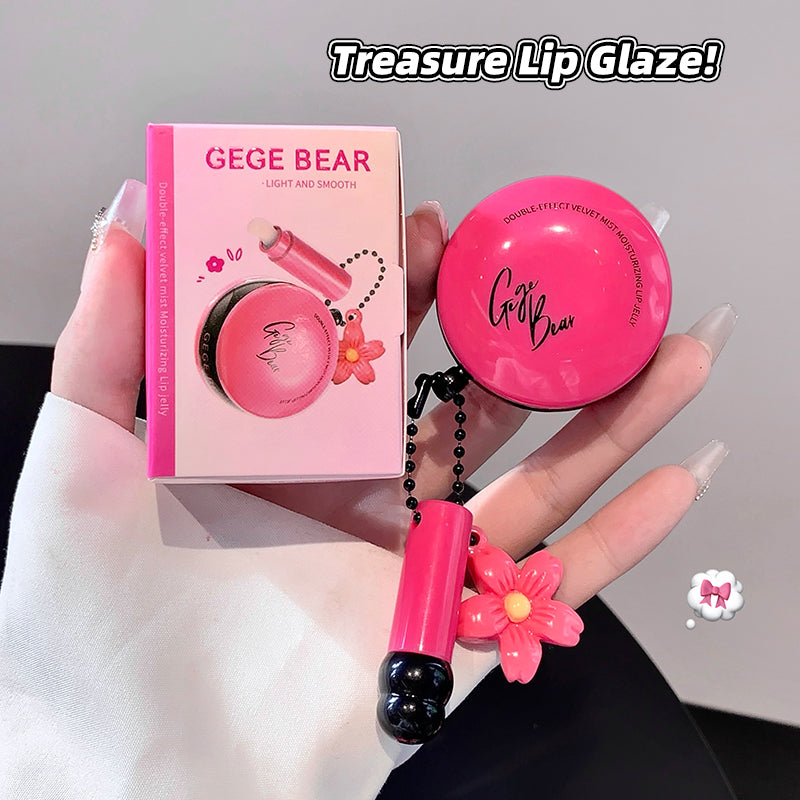 Pink lip gloss with floral design held by a hand, next to its packaging, on a neutral background.