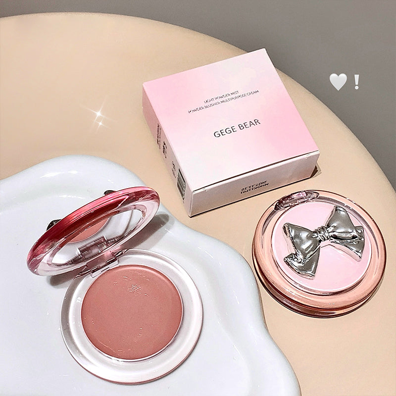 Makeup compact with pink blush and bow design, accompanied by a pink box labeled 'GEGE BEAR' on a beige surface.