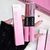 Two GEUGE BEAR lip glosses held in front of their packaging on a white background.