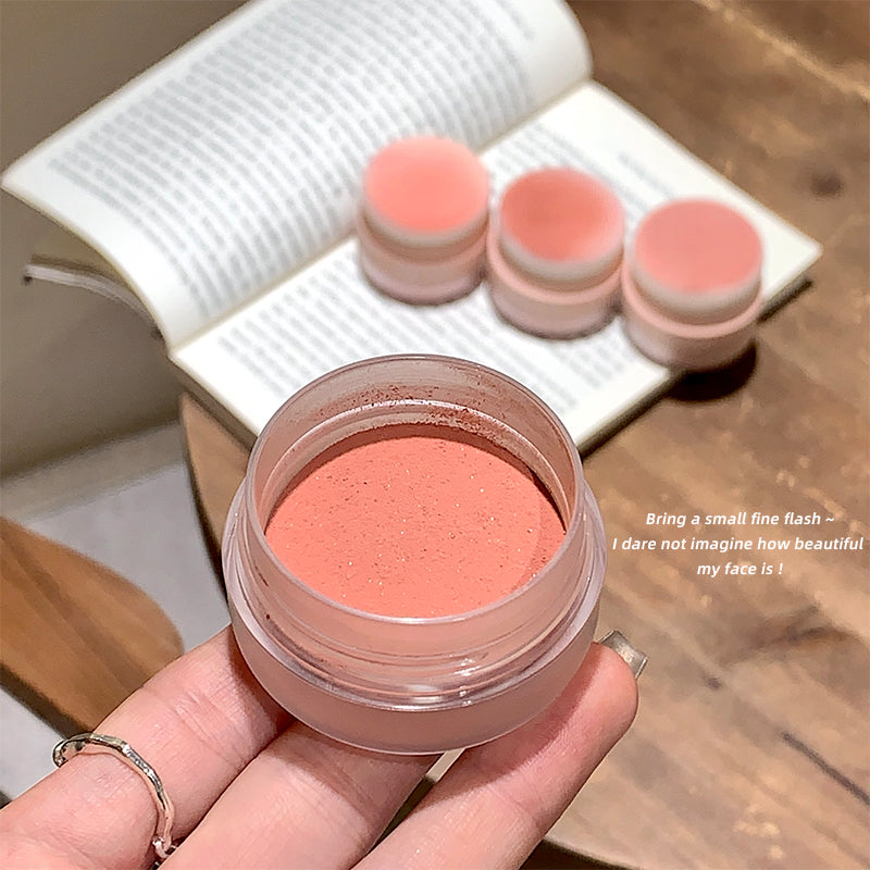 Hand holding a jar of pink cream with three other jars in the background on a wooden surface.
