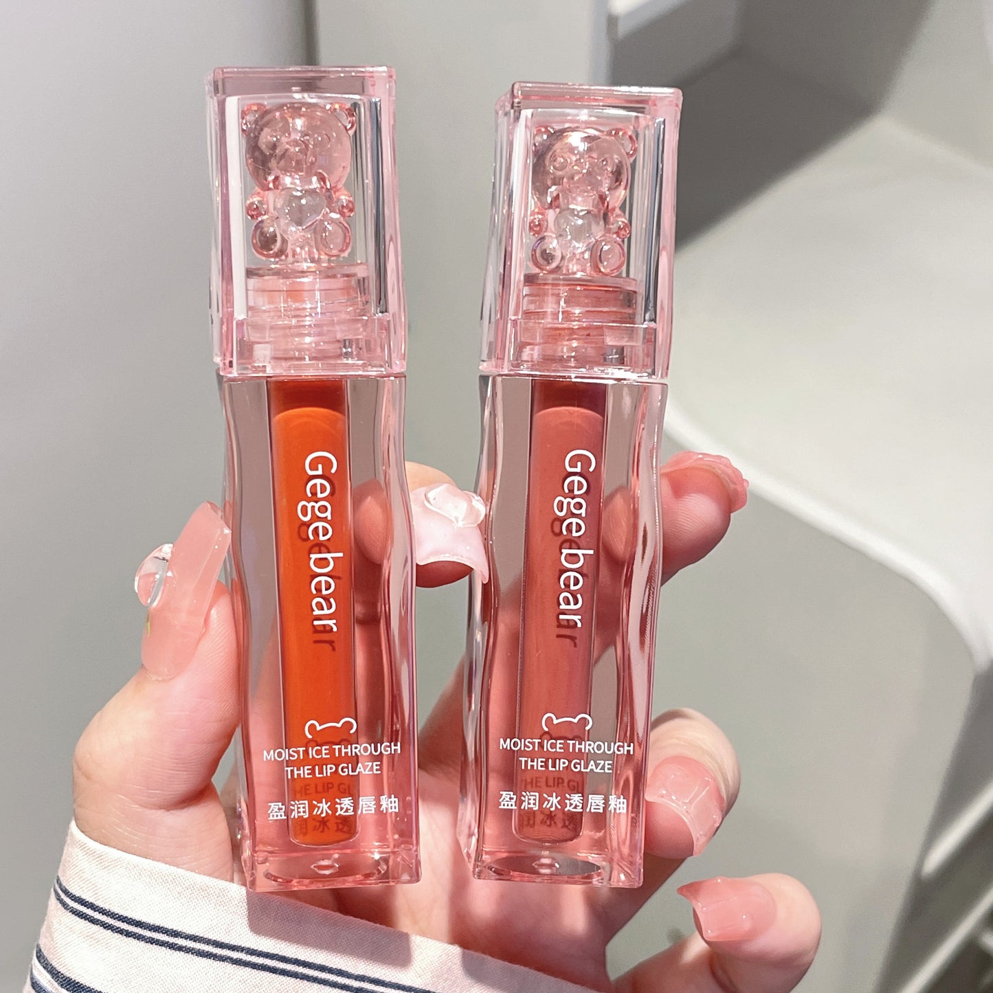 Two lip glosses with 'Gege bear' branding held in a hand against a neutral background