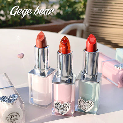 Three lipsticks with heart-shaped caps on a reflective surface, with 'Gege bear' branding.