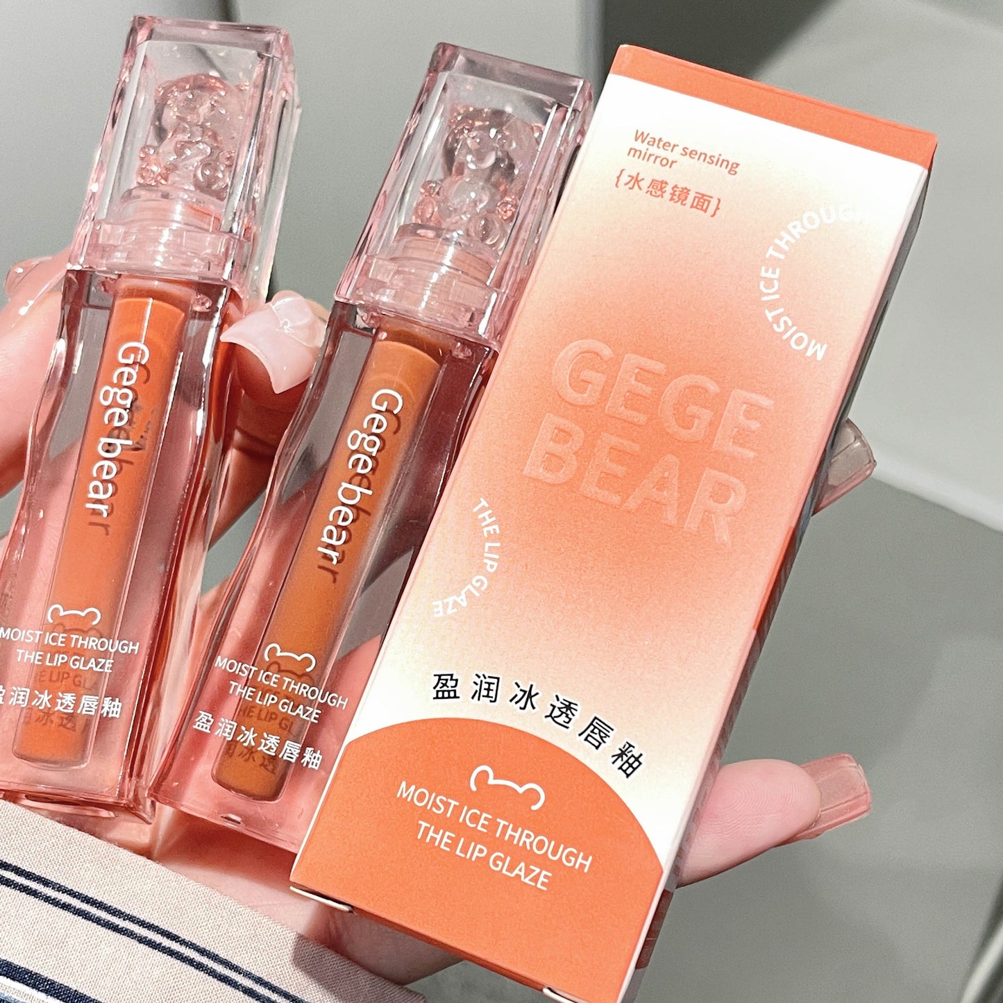 Two lip glosses and their packaging held by a hand on a neutral background