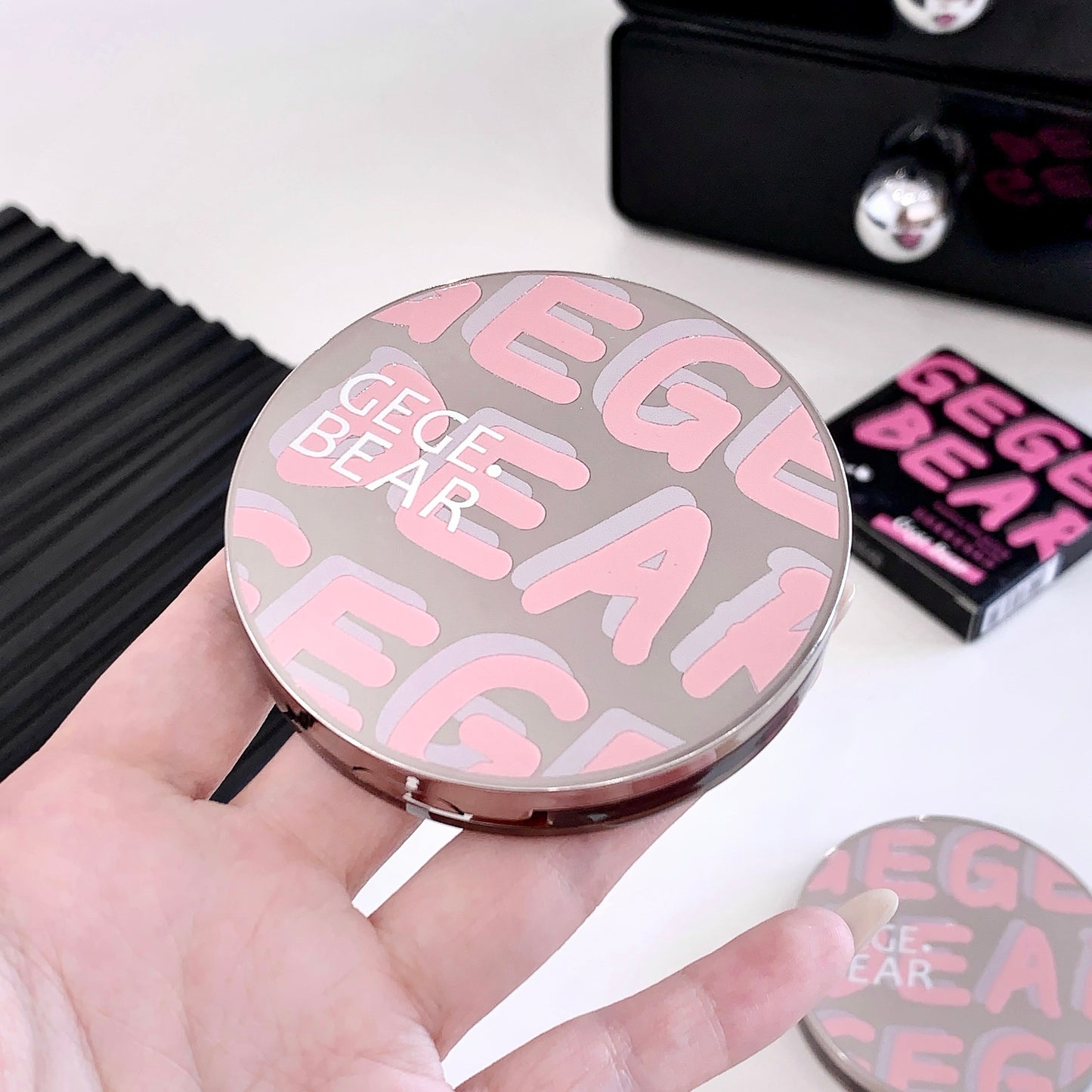 Compact mirror with 'GEGE BEAR' branding held in a hand, with a blurred background.