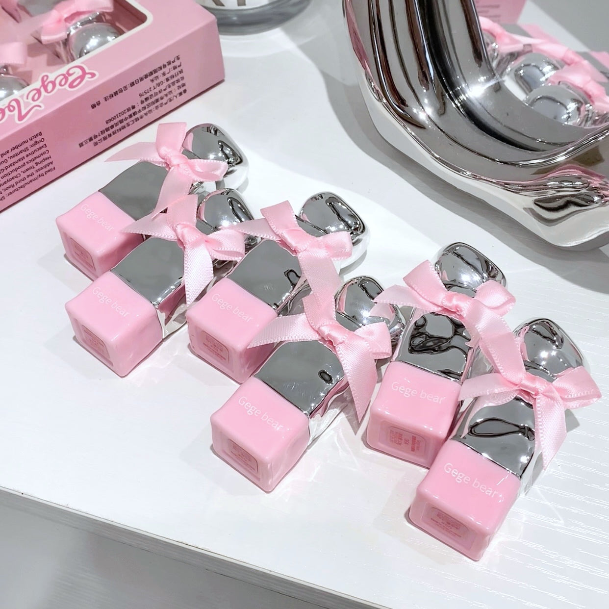 Pink and silver products with ribbons on a white surface, next to a pink box.