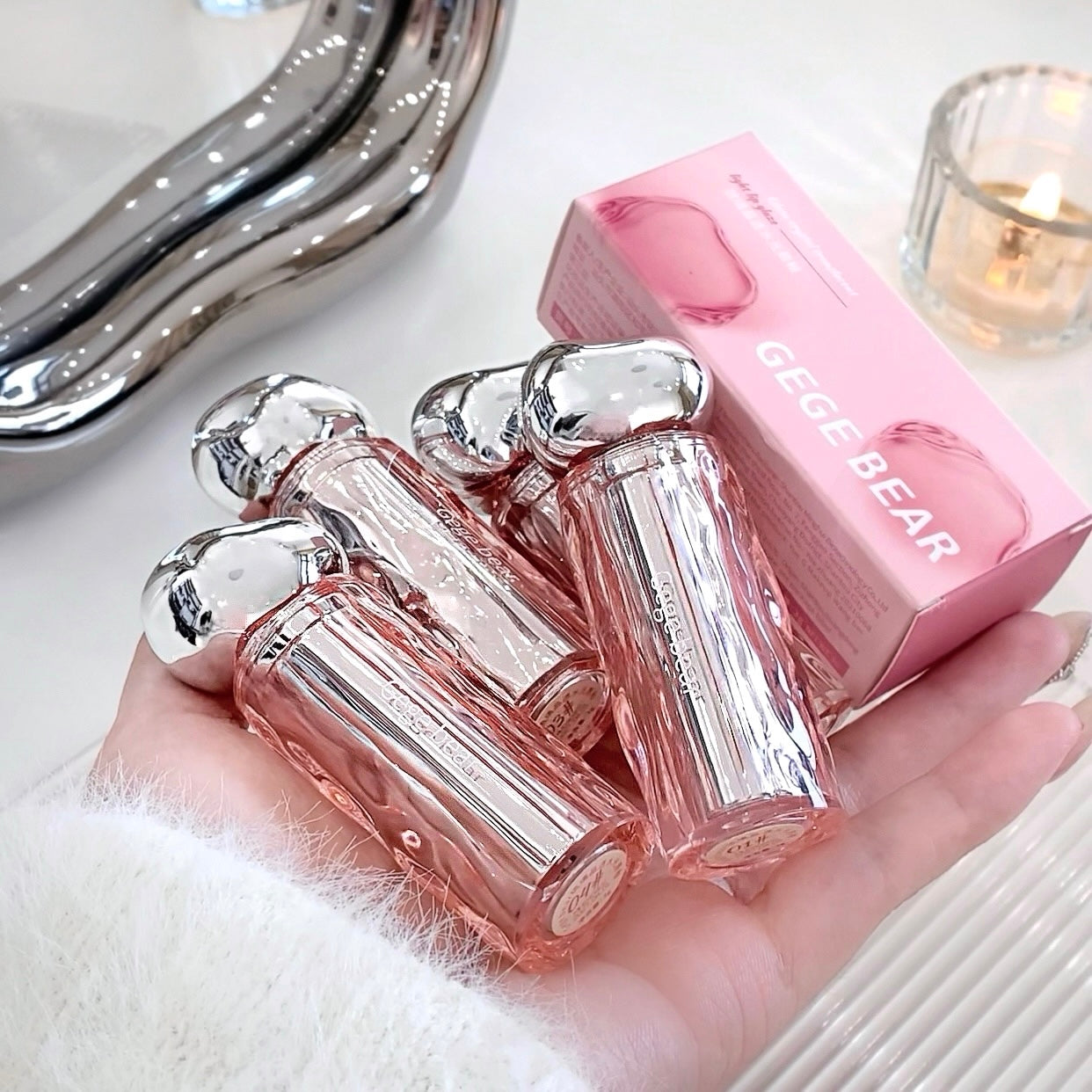 Pink lip glosses with silver caps on a hand, next to a pink 'GEGE BEAR' box.