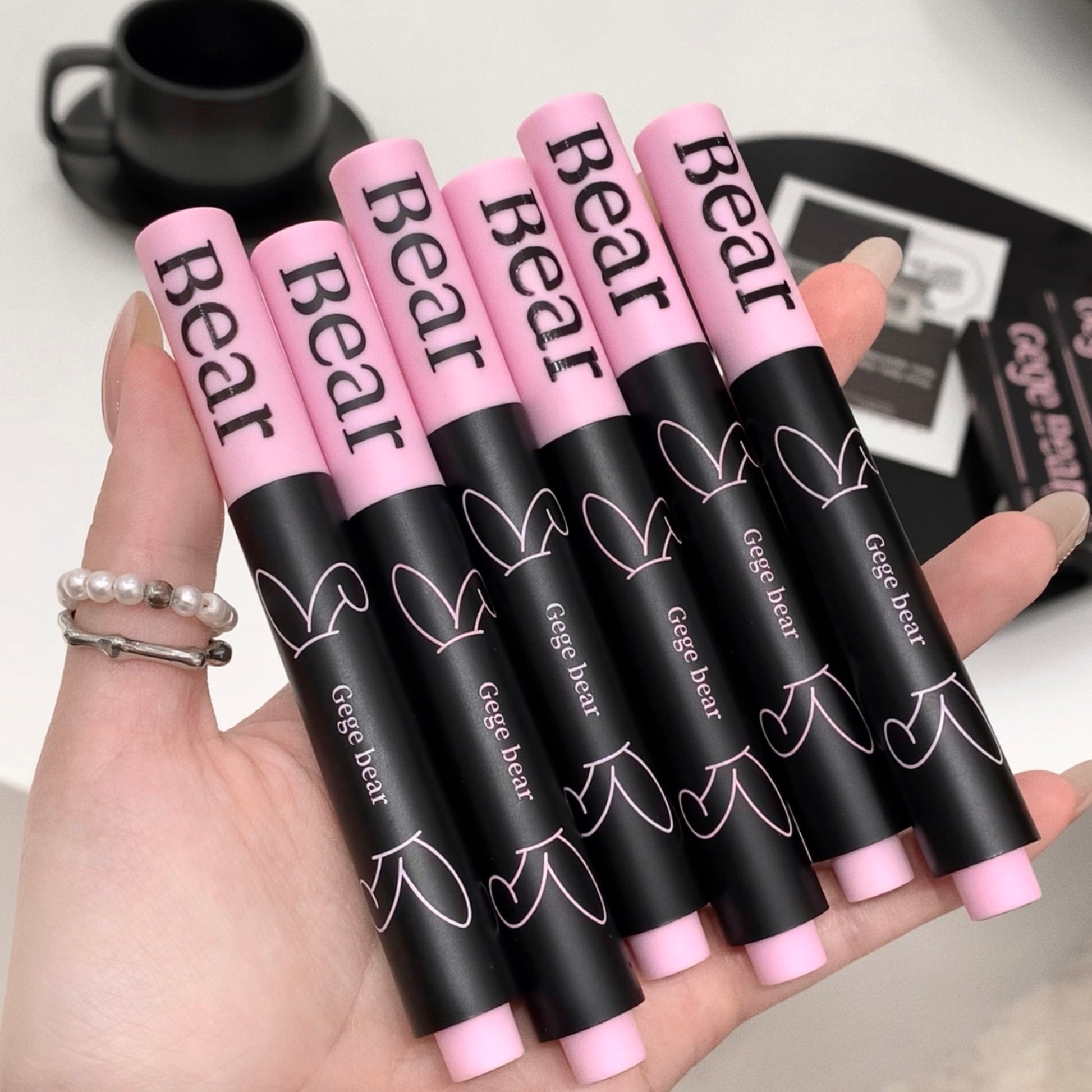 Hand holding six black and pink 'Bear' lip glosses with a blurred background