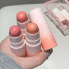 Three blush sticks with a pink container held in a hand on a light surface.