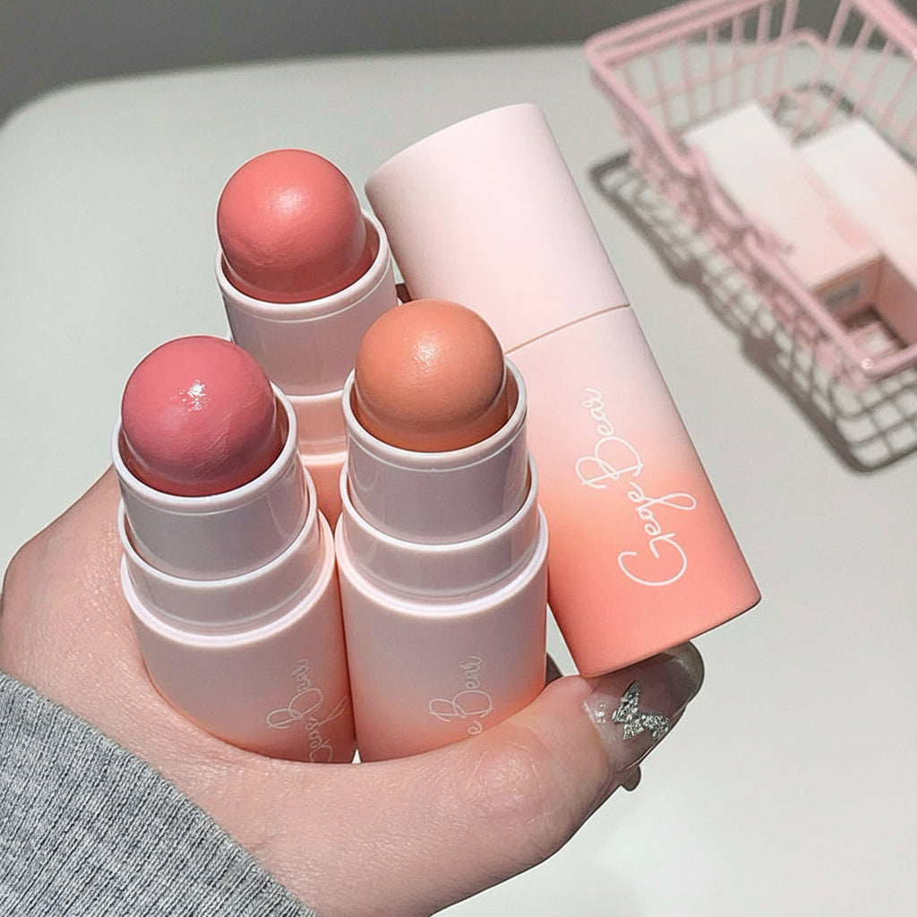 Three blush sticks with a pink container held in a hand on a light surface.