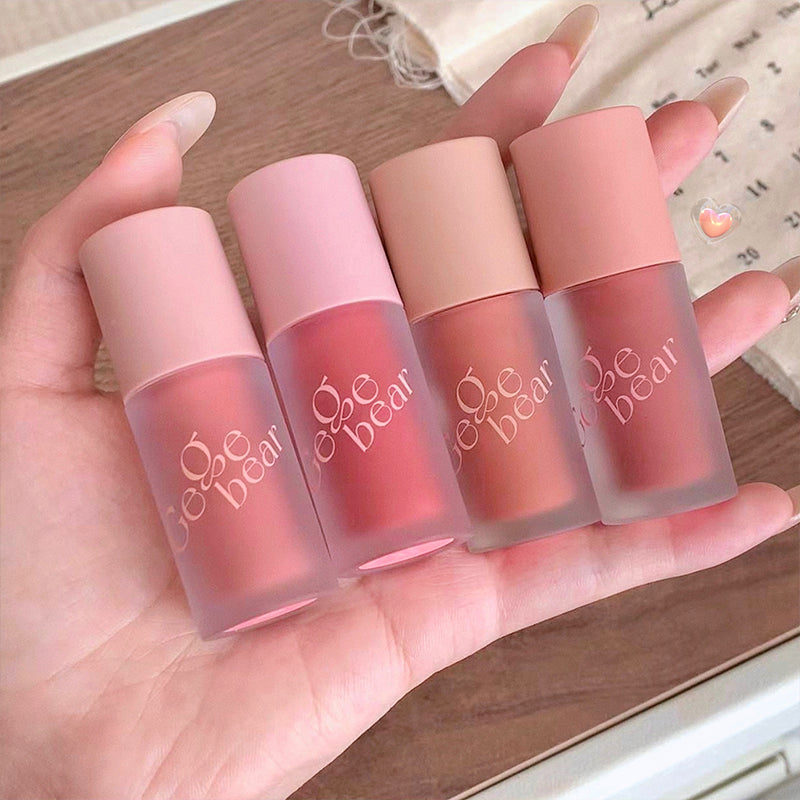 Four lip glosses from the brand 'be bear' held in a hand with a blurred background.