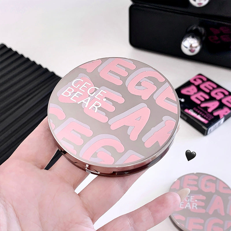 Compact mirror with 'GEGE BEAR' branding held in a hand on a blurred background.